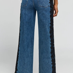 Back view of a model wearing blue wide-leg denim jeans with black lace trim down the outer sides, paired with a navy top and black heels, against a light gray studio background.