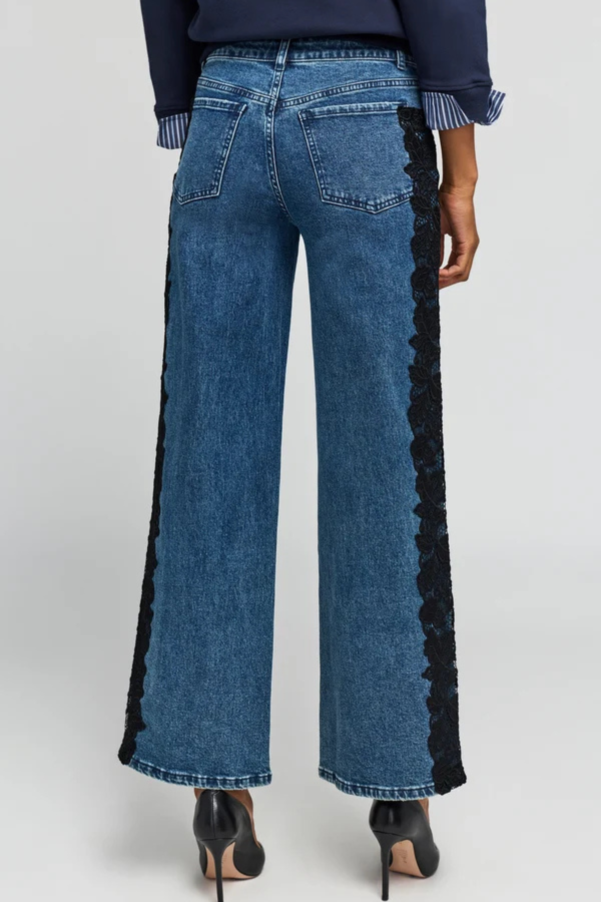 Back view of a model wearing blue wide-leg denim jeans with black lace trim down the outer sides, paired with a navy top and black heels, against a light gray studio background.