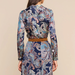 A woman shown from the back wearing a long-sleeve collared dress with a blue, beige, and brown paisley pattern and a tan belt, against a solid beige background.