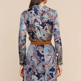 A woman shown from the back wearing a long-sleeve collared dress with a blue, beige, and brown paisley pattern and a tan belt, against a solid beige background.