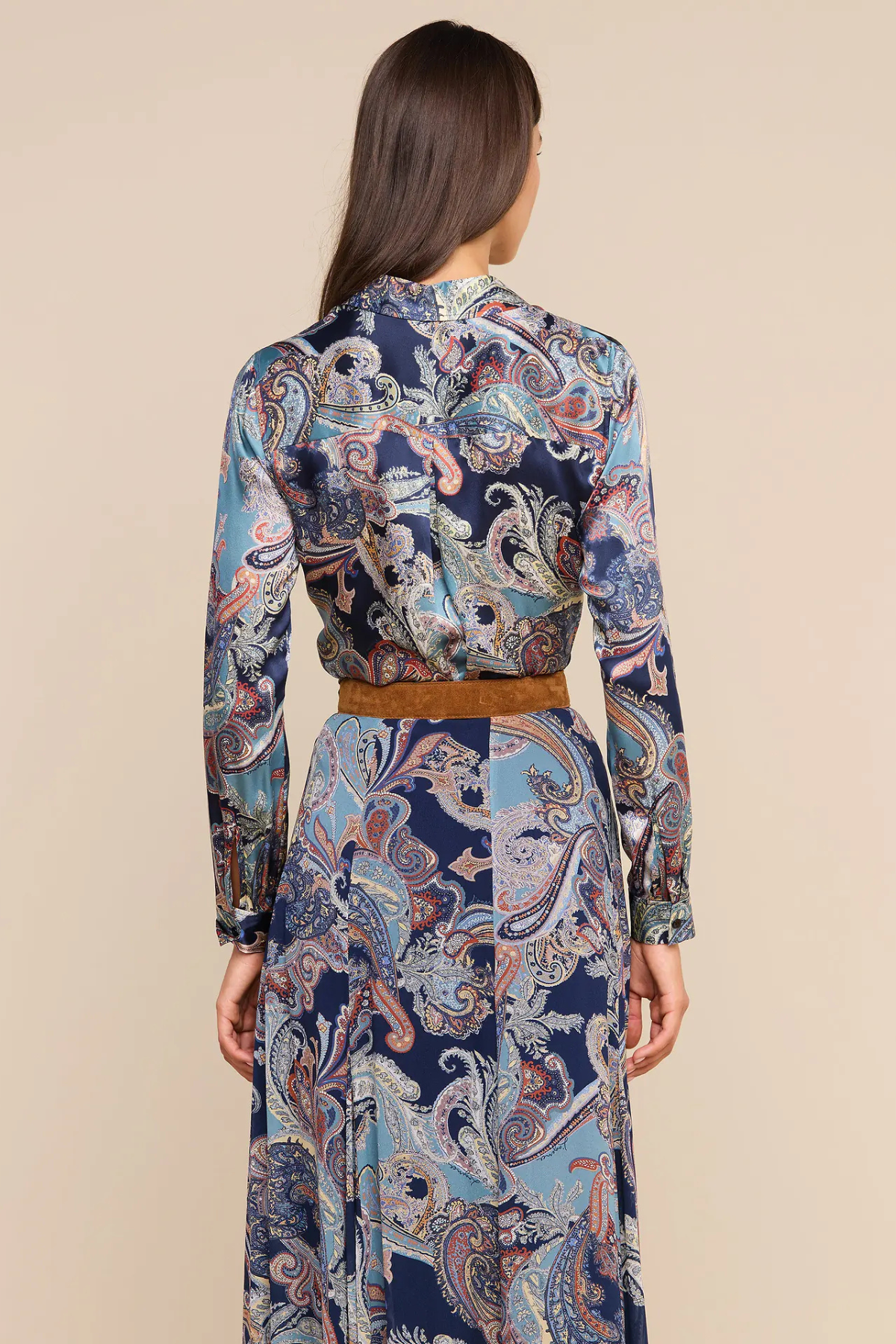 A woman shown from the back wearing a long-sleeve collared dress with a blue, beige, and brown paisley pattern and a tan belt, against a solid beige background.