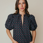 A woman modeling a short-sleeve blouse with puff sleeves and a split V-neckline, featuring a dark navy base with a small multicolor geometric pattern, paired with light blue jeans against a solid beige background.