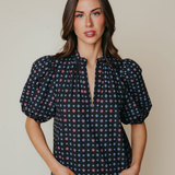 A woman modeling a short-sleeve blouse with puff sleeves and a split V-neckline, featuring a dark navy base with a small multicolor geometric pattern, paired with light blue jeans against a solid beige background.