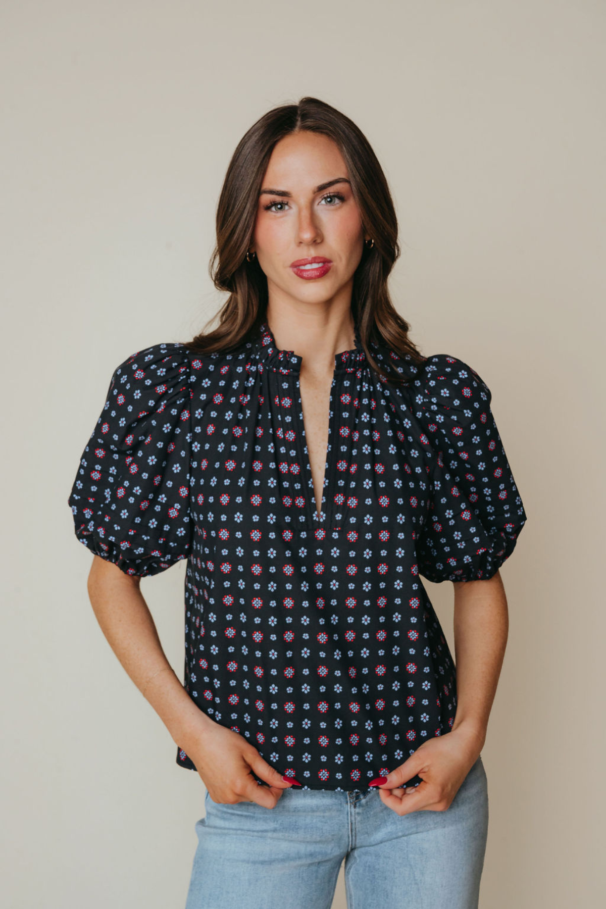 A woman modeling a short-sleeve blouse with puff sleeves and a split V-neckline, featuring a dark navy base with a small multicolor geometric pattern, paired with light blue jeans against a solid beige background.