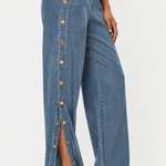 A person wearing high-waisted wide-leg denim pants with a vertical row of gold-tone buttons down the front slit, paired with metallic heeled sandals against a plain light background.