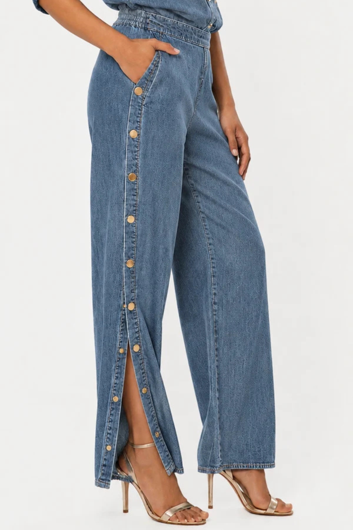 A person wearing high-waisted wide-leg denim pants with a vertical row of gold-tone buttons down the front slit, paired with metallic heeled sandals against a plain light background.