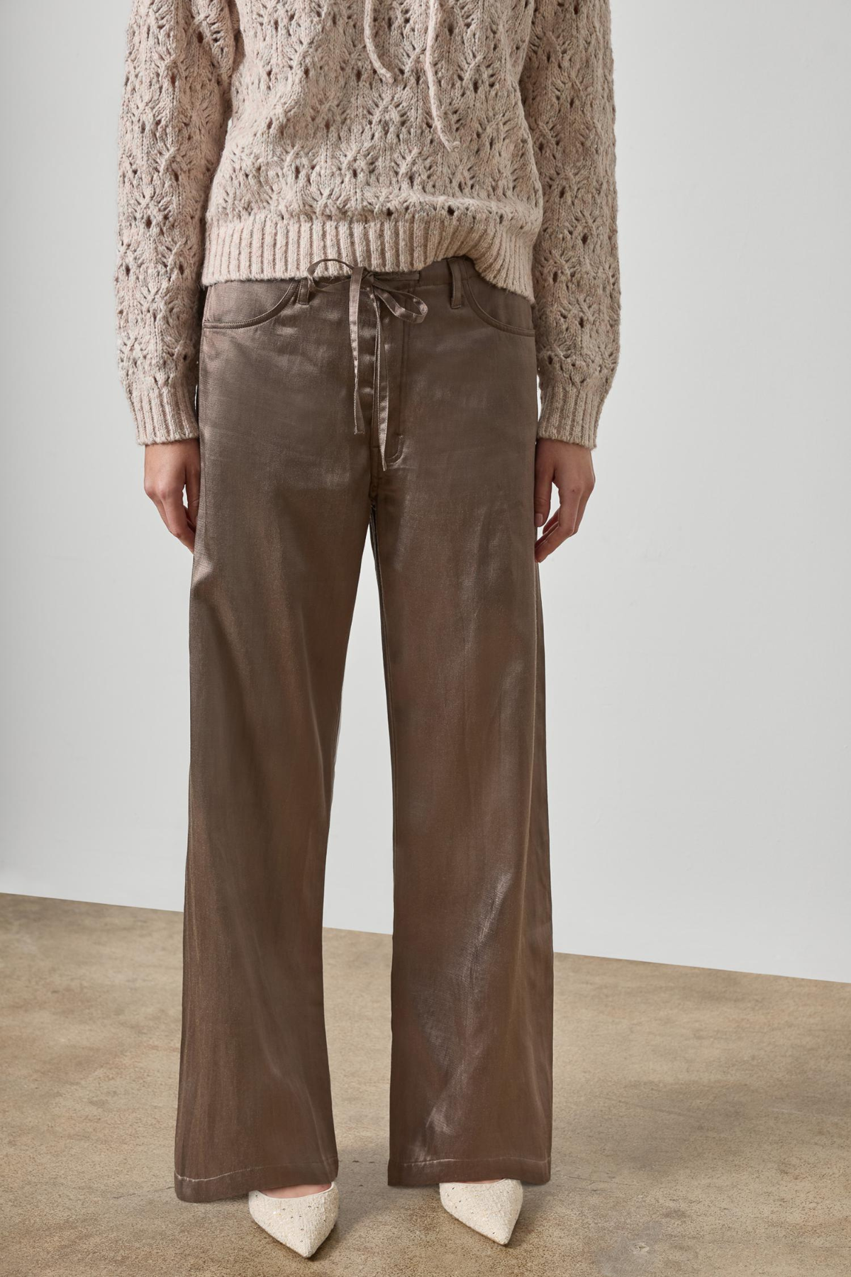 A model wearing a beige cable-knit sweater and brown wide-leg pants with a satin-like sheen, paired with white pointed-toe heels, set against a minimalist studio background.