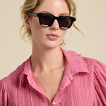 Woman wearing black sunglasses and a pink checkered shirt against a beige background