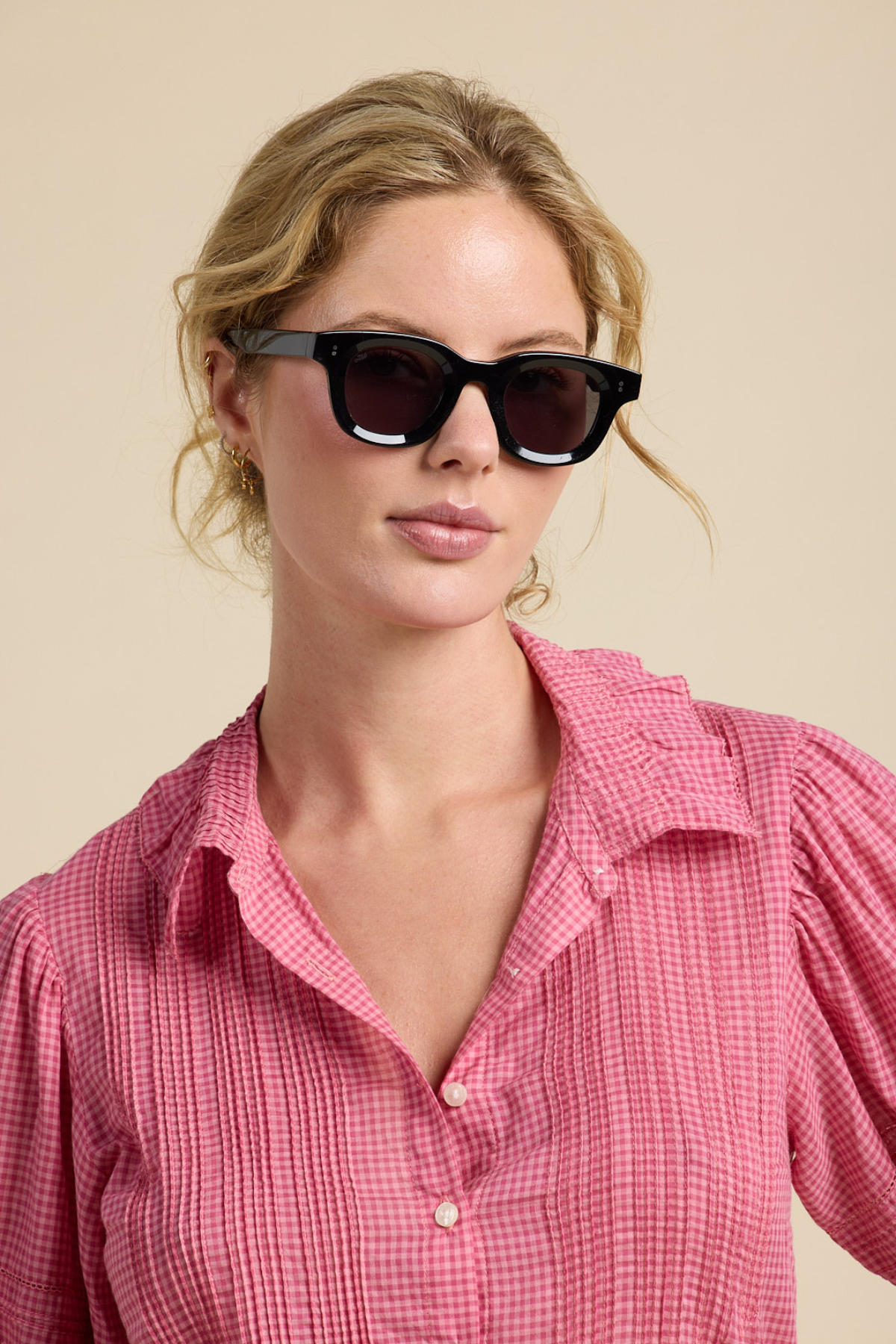 Woman wearing black sunglasses and a pink checkered shirt against a beige background