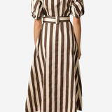 A woman viewed from the back wearing a midi dress with vertical brown and cream stripes, short puff sleeves, and a matching belt, against a plain light gray background.