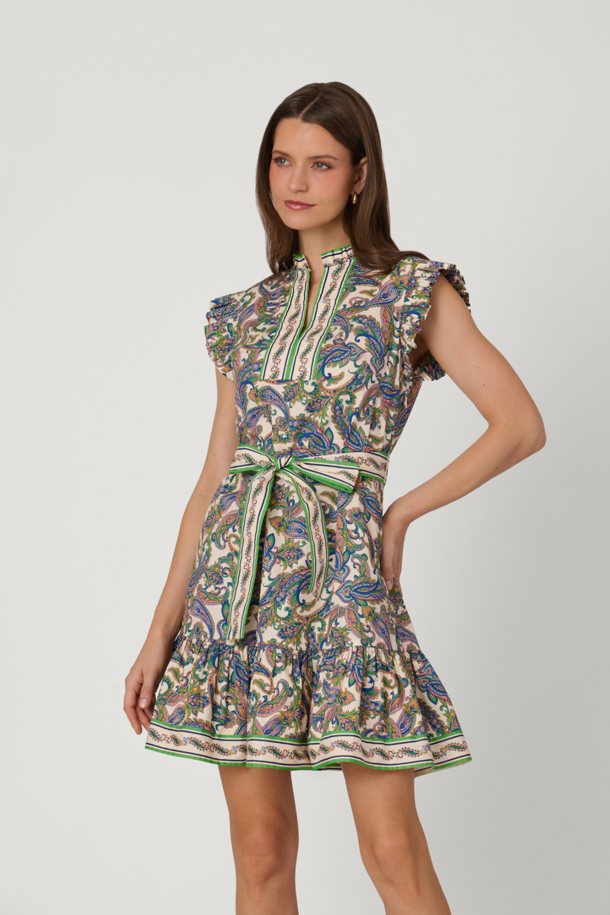A woman modeling a short, sleeveless dress with a high mock neck, ruffled cap sleeves, and a matching tie belt at the waist. The dress features an intricate multicolor paisley pattern with green, blue, beige, and brown tones, set against a plain light gray studio background.
