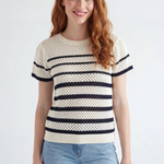 A woman with long red hair wearing a cream-colored knit short-sleeve sweater with horizontal black stripes and light blue jeans, standing against a light gray studio background.