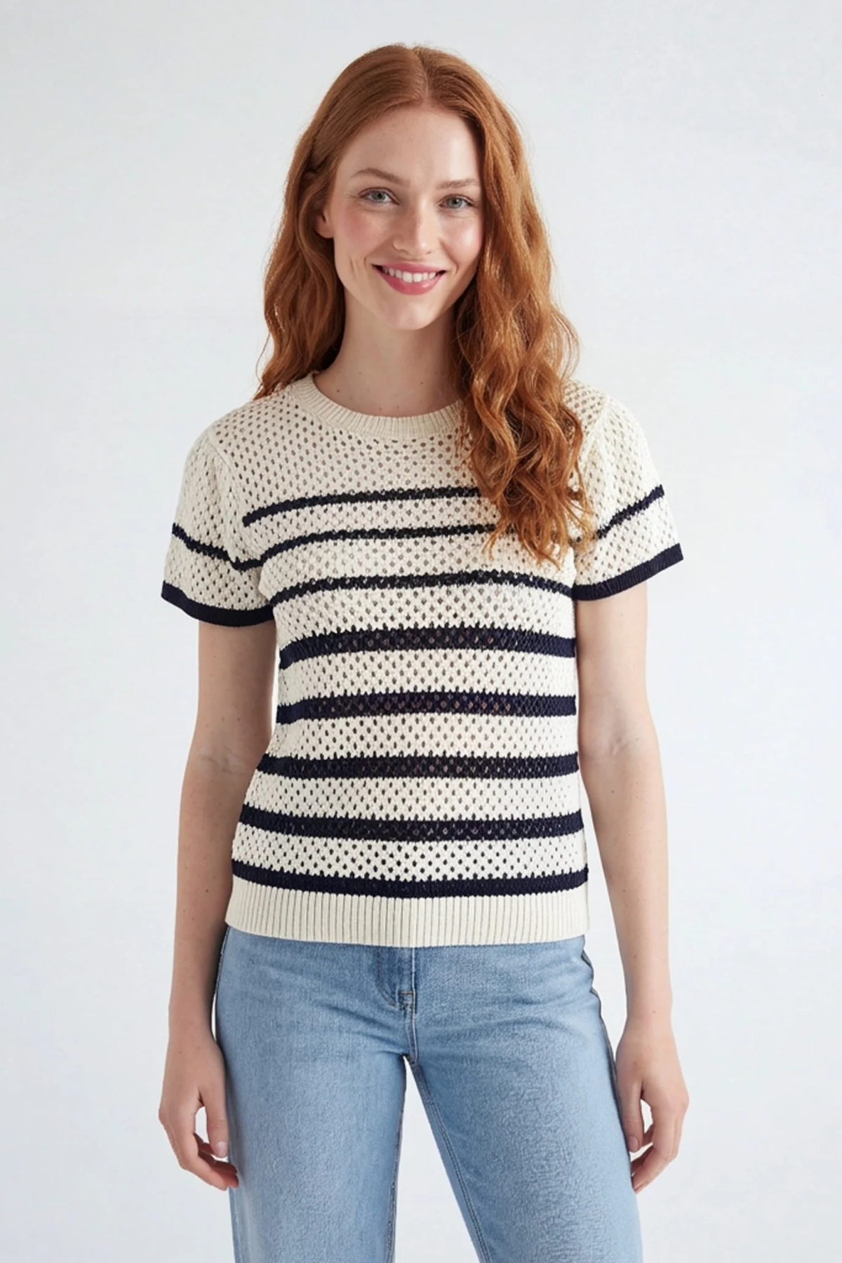 A woman with long red hair wearing a cream-colored knit short-sleeve sweater with horizontal black stripes and light blue jeans, standing against a light gray studio background.