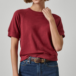 A woman wearing a short-sleeve crewneck t-shirt in a heathered burgundy red, paired with dark wash blue jeans and a brown belt, photographed against a neutral light gray background.