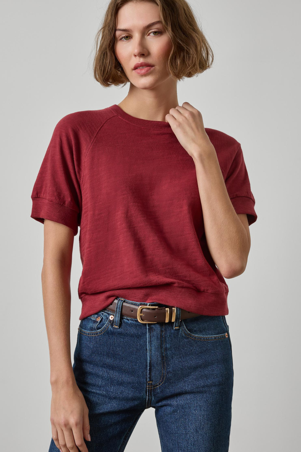 A woman wearing a short-sleeve crewneck t-shirt in a heathered burgundy red, paired with dark wash blue jeans and a brown belt, photographed against a neutral light gray background.