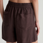 A close-up back view of a model wearing dark brown linen shorts with an elastic waistband and side pockets, set against a light gray studio background.