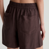 A close-up back view of a model wearing dark brown linen shorts with an elastic waistband and side pockets, set against a light gray studio background.