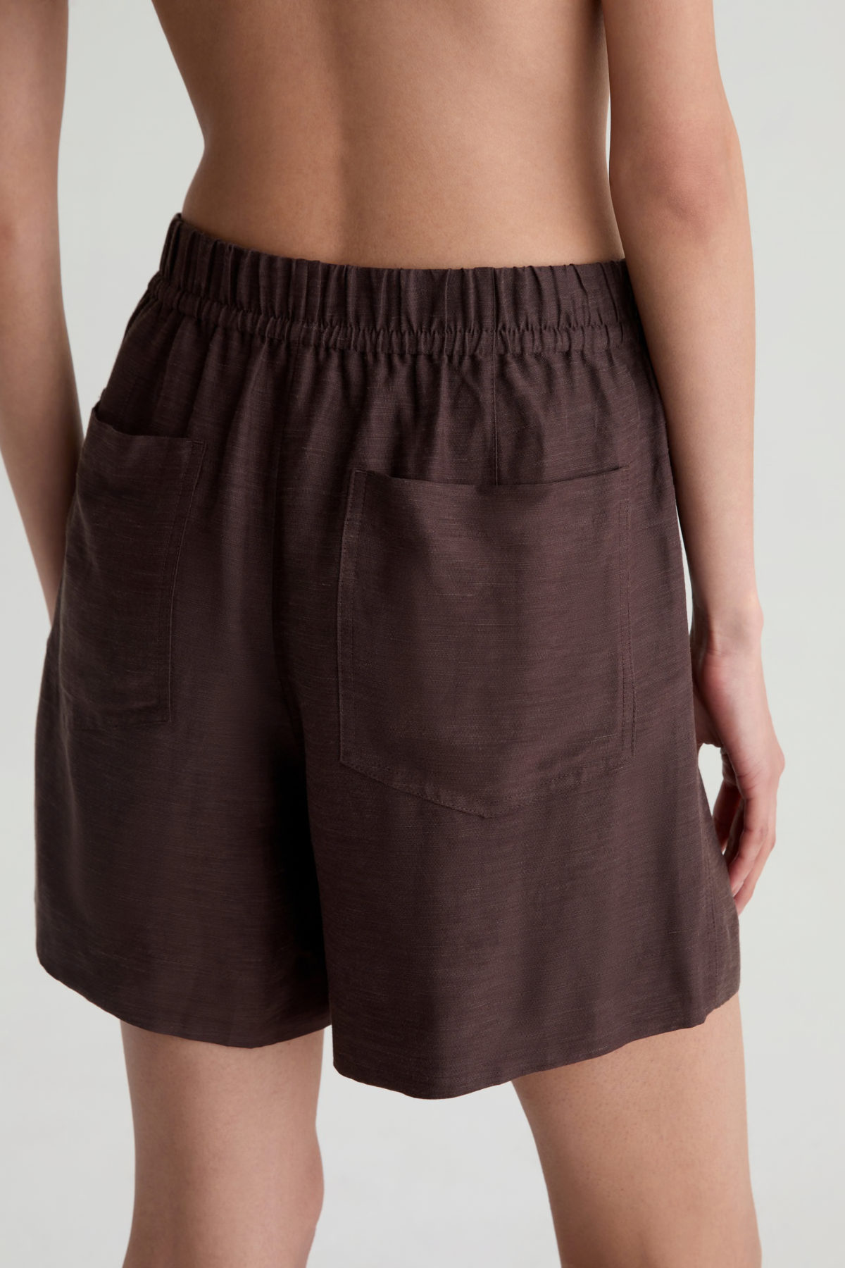 A close-up back view of a model wearing dark brown linen shorts with an elastic waistband and side pockets, set against a light gray studio background.