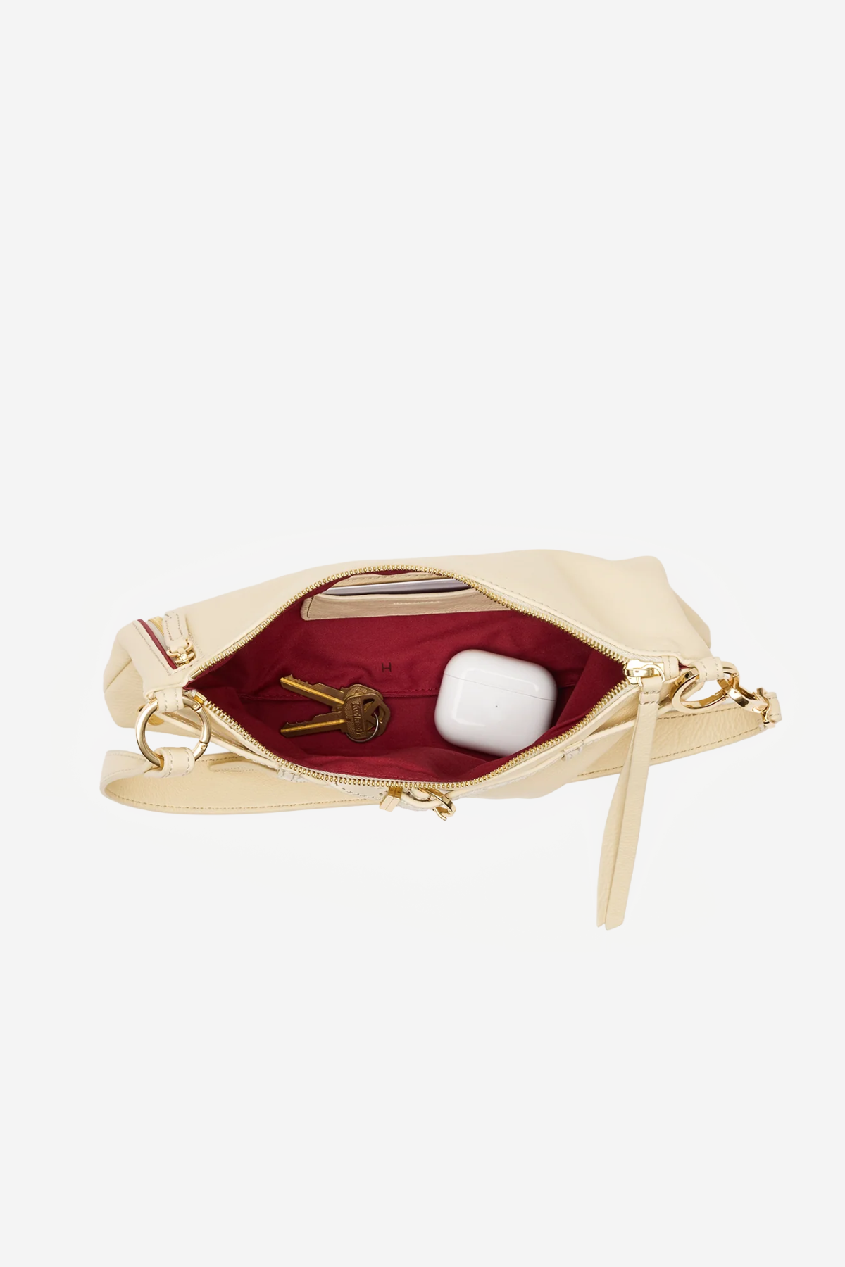 An open cream-colored leather clutch bag with gold-tone hardware, showing a deep burgundy red interior lining. Inside, a gold key and a white wireless earbud case are visible.
