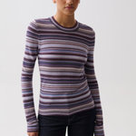 A woman modeling a long-sleeve crewneck knit top with horizontal stripes in purple, blue, pink, and beige, paired with dark jeans against a light gray studio background.