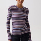 A woman modeling a long-sleeve crewneck knit top with horizontal stripes in purple, blue, pink, and beige, paired with dark jeans against a light gray studio background.