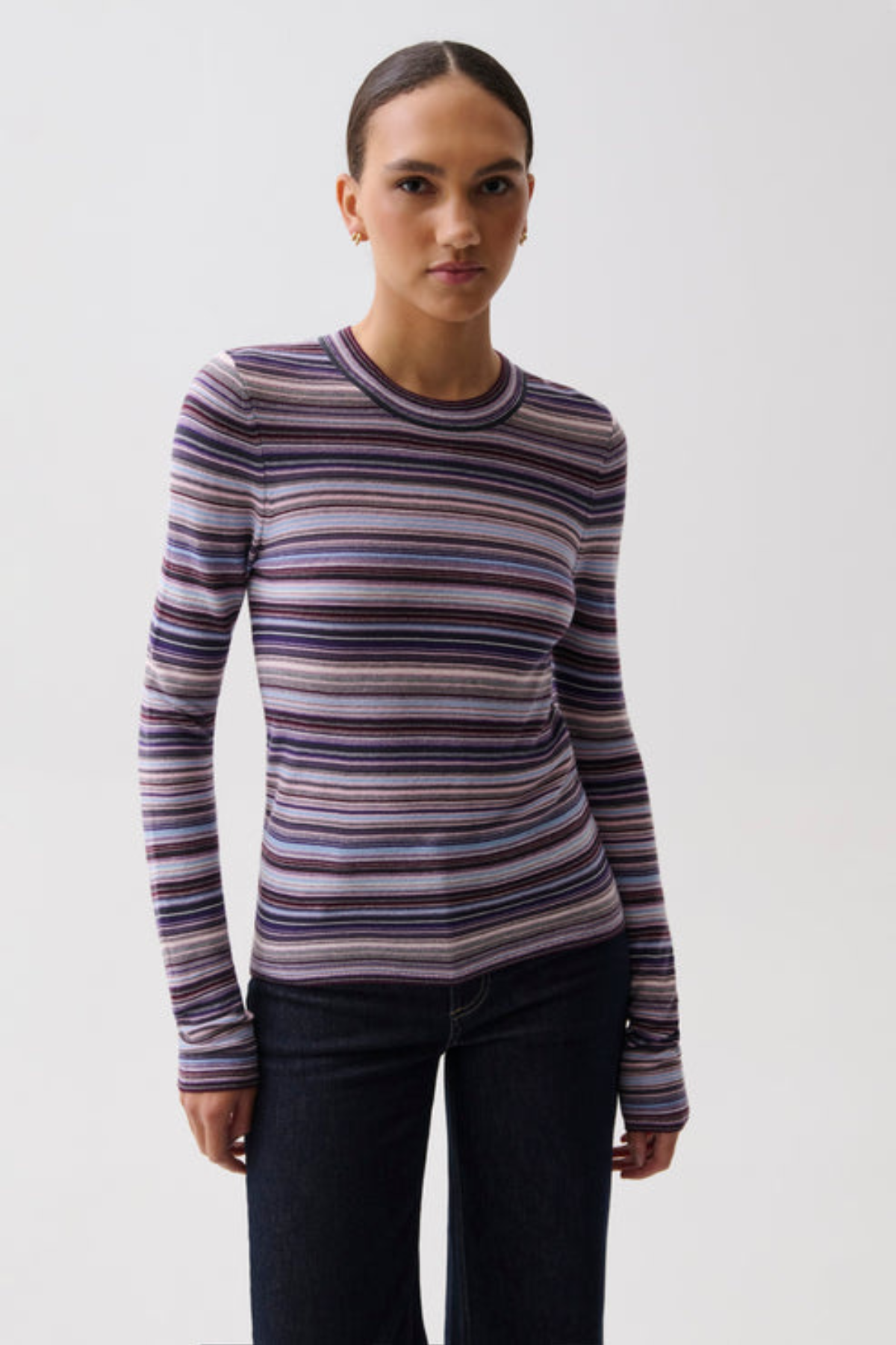 A woman modeling a long-sleeve crewneck knit top with horizontal stripes in purple, blue, pink, and beige, paired with dark jeans against a light gray studio background.
