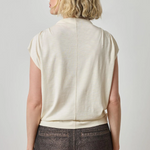 Woman wearing a beige sleeveless top and dark pants against a plain background