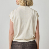 Woman wearing a beige sleeveless top and dark pants against a plain background