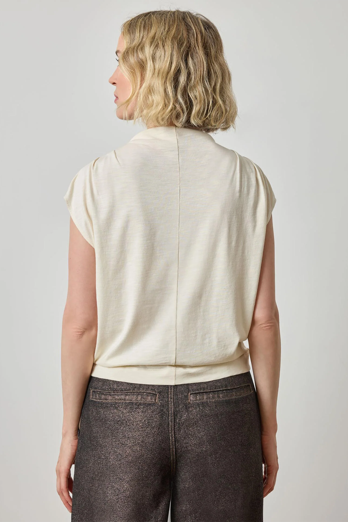 Woman wearing a beige sleeveless top and dark pants against a plain background