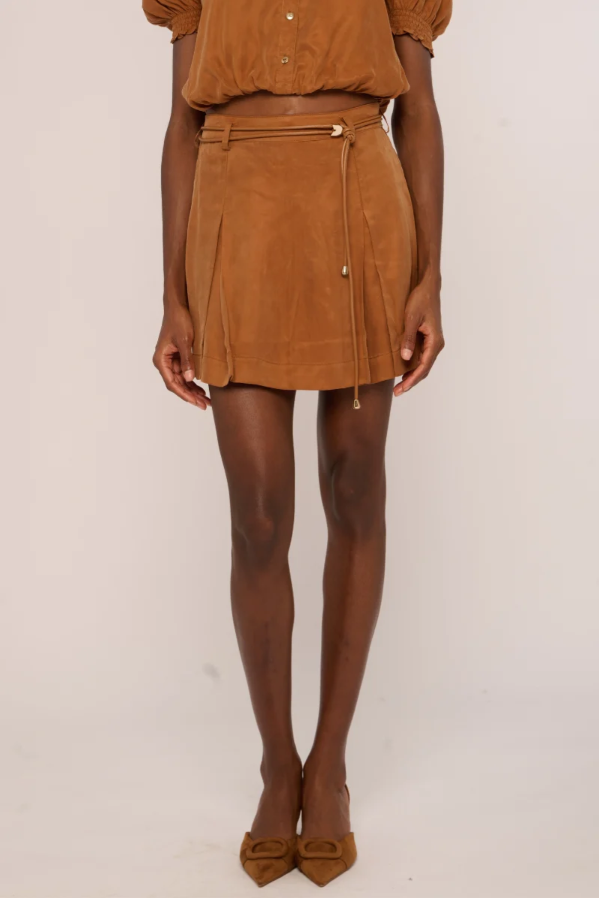 A model wearing a brown suede mini skirt with a matching belt and tie, paired with a matching brown top and brown pointed-toe flats, against a plain light background.