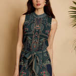 A woman modeling a sleeveless high-neck jumpsuit with a dark teal and navy geometric pattern, floral accents, and a matching waist tie, set against a warm beige background with a plant leaf on the right.
