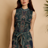 A woman modeling a sleeveless high-neck jumpsuit with a dark teal and navy geometric pattern, floral accents, and a matching waist tie, set against a warm beige background with a plant leaf on the right.