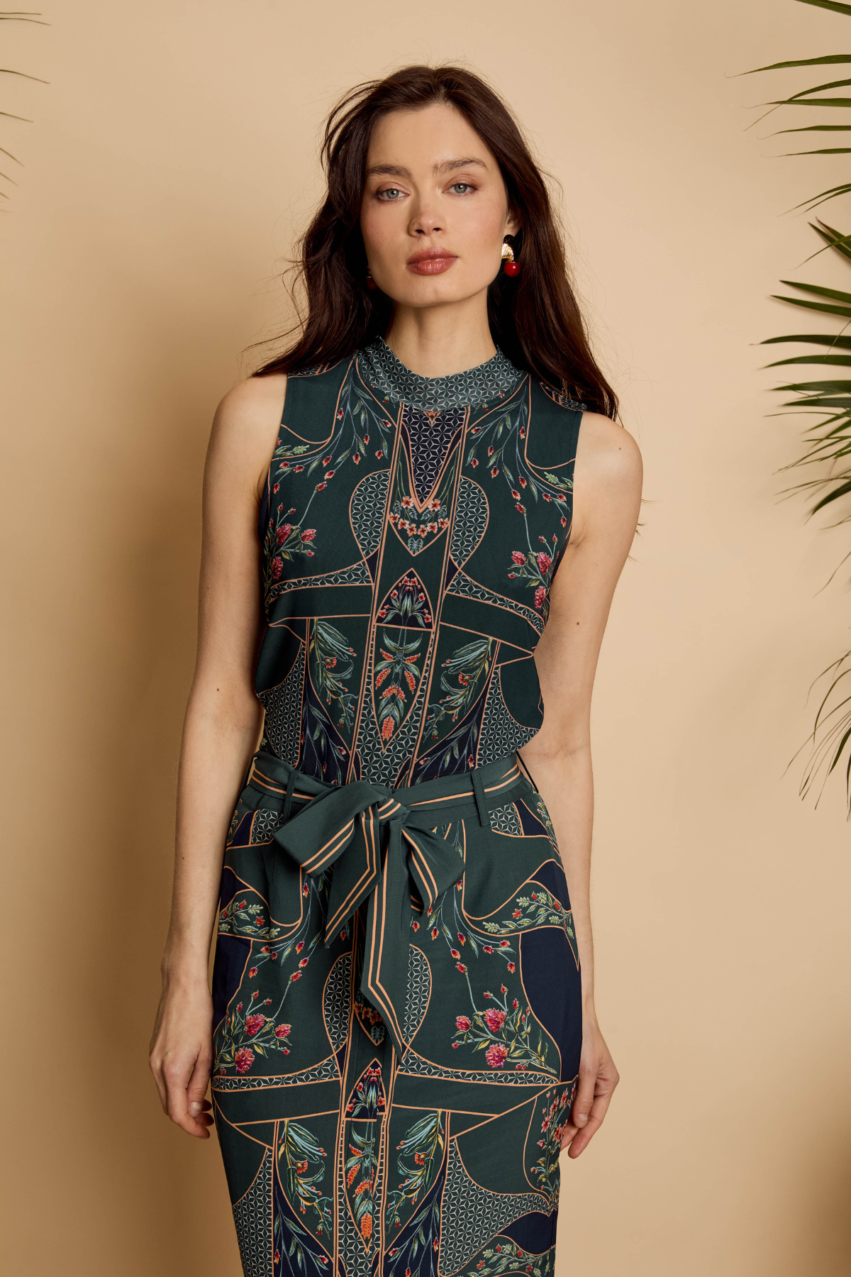 A woman modeling a sleeveless high-neck jumpsuit with a dark teal and navy geometric pattern, floral accents, and a matching waist tie, set against a warm beige background with a plant leaf on the right.