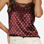 A woman wearing a burgundy satin camisole top with white polka dots and a dark lace neckline, paired with off-white pants, against a plain light background.