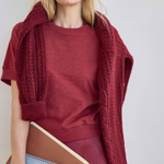 A woman wearing a rust-red crewneck top, matching skirt, and a chunky cable-knit shrug draped over her shoulders, holding brown and blue notebooks against a light gray background.