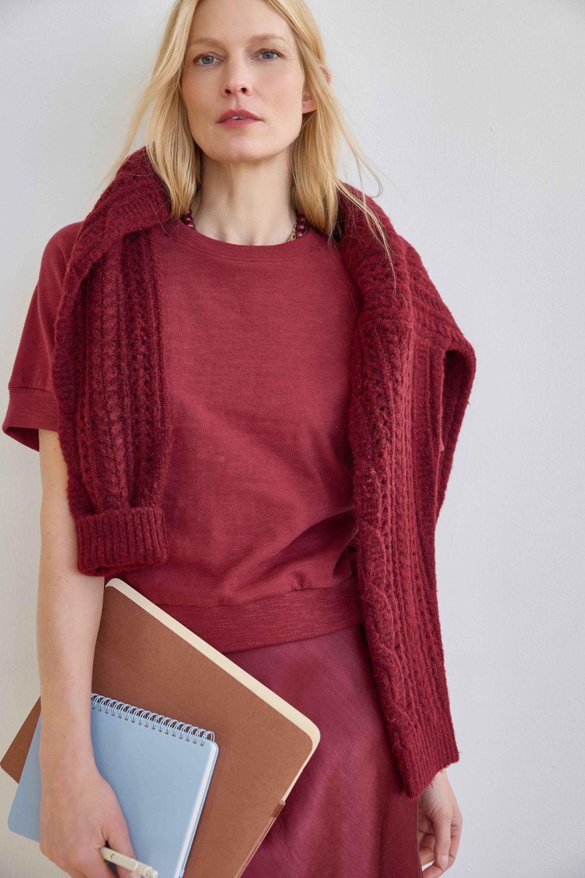 A woman wearing a rust-red crewneck top, matching skirt, and a chunky cable-knit shrug draped over her shoulders, holding brown and blue notebooks against a light gray background.