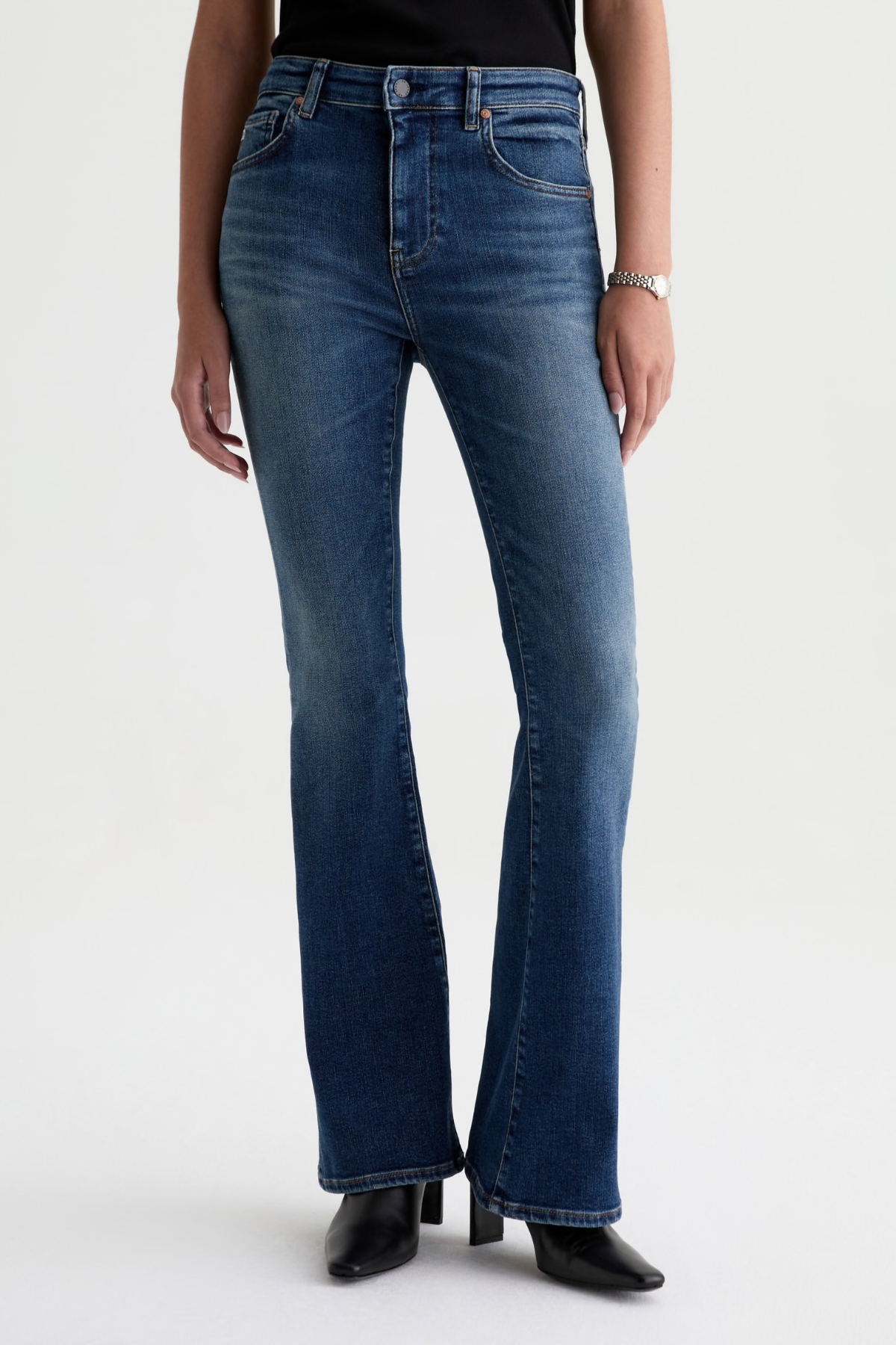A model wearing a pair of dark blue high-waisted flare jeans with a subtle whiskering effect at the thighs, paired with a black top and black pointed-toe boots against a light gray studio background.