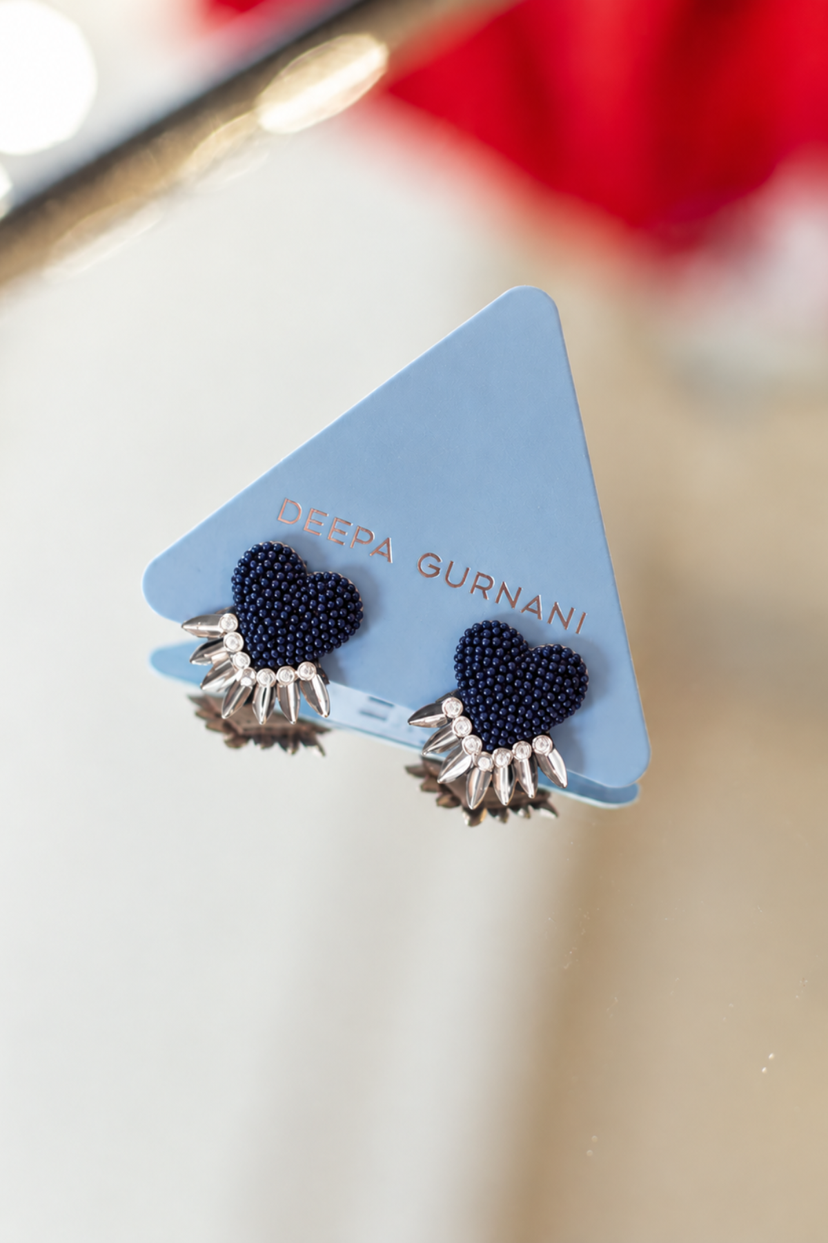 A pair of Deepa Gurnani stud earrings displayed on a light blue triangular card. The earrings feature dark navy blue seed bead hearts with silver-toned metal spikes extending from the bottom.