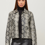 A woman modeling a cropped long-sleeve jacket with a gray and black snakeskin pattern, worn over a black turtleneck and black faux-leather pants, against a plain light gray studio background.
