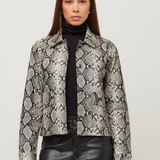 A woman modeling a cropped long-sleeve jacket with a gray and black snakeskin pattern, worn over a black turtleneck and black faux-leather pants, against a plain light gray studio background.