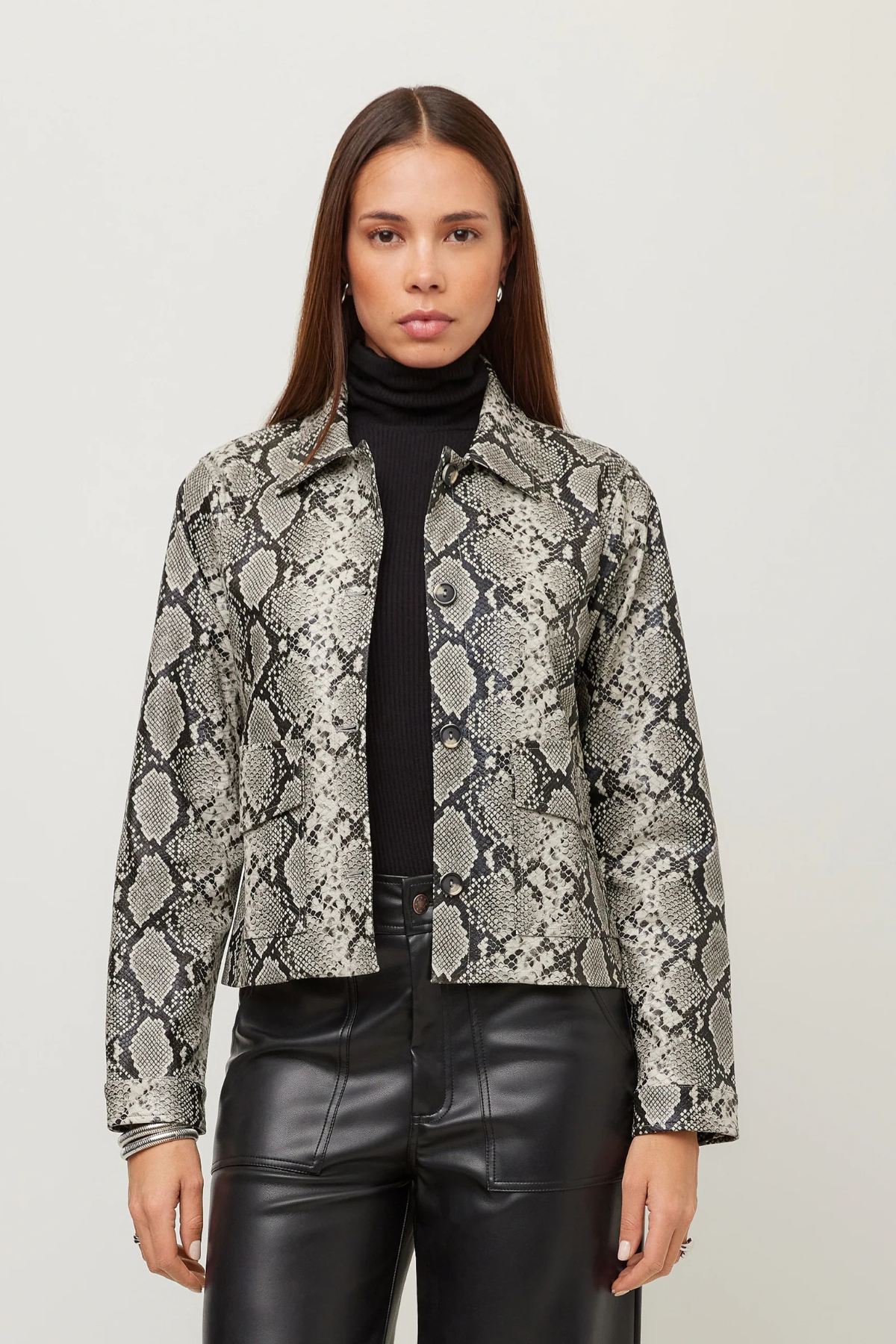A woman modeling a cropped long-sleeve jacket with a gray and black snakeskin pattern, worn over a black turtleneck and black faux-leather pants, against a plain light gray studio background.