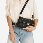 A model wearing a cream satin shirt and light-wash jeans carries a black leather crossbody bag with gold-tone hardware and studs against a plain white background.