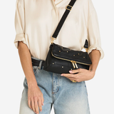 A model wearing a cream satin shirt and light-wash jeans carries a black leather crossbody bag with gold-tone hardware and studs against a plain white background.