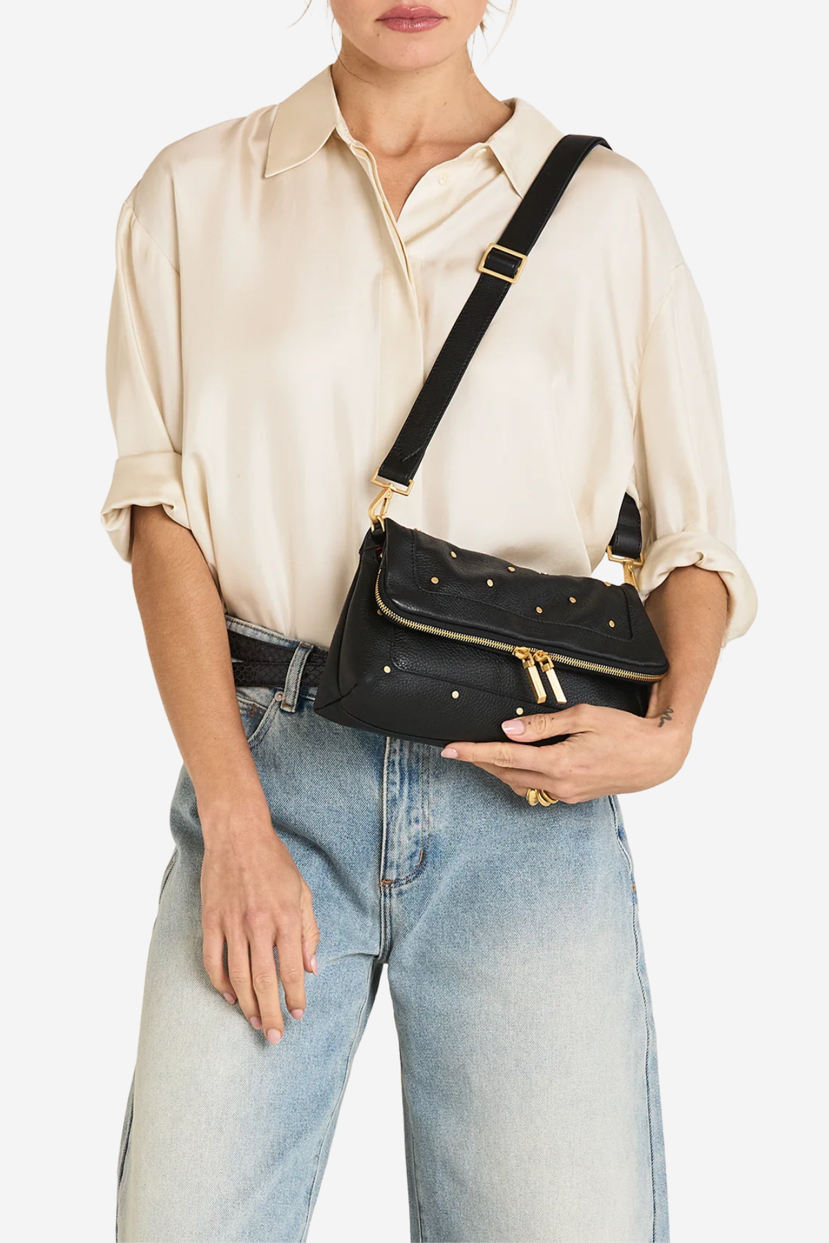 A model wearing a cream satin shirt and light-wash jeans carries a black leather crossbody bag with gold-tone hardware and studs against a plain white background.