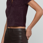 A close-up of a model wearing a short-sleeve fuzzy knit polo crop top in deep plum purple, paired with dark brown faux-leather pants, against a light gray background.