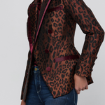 A woman wearing a fitted leopard-print blazer with dark burgundy satin lapels and buttons, paired with blue jeans, photographed against a light gray studio background.