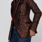 A woman wearing a fitted leopard-print blazer with dark burgundy satin lapels and buttons, paired with blue jeans, photographed against a light gray studio background.