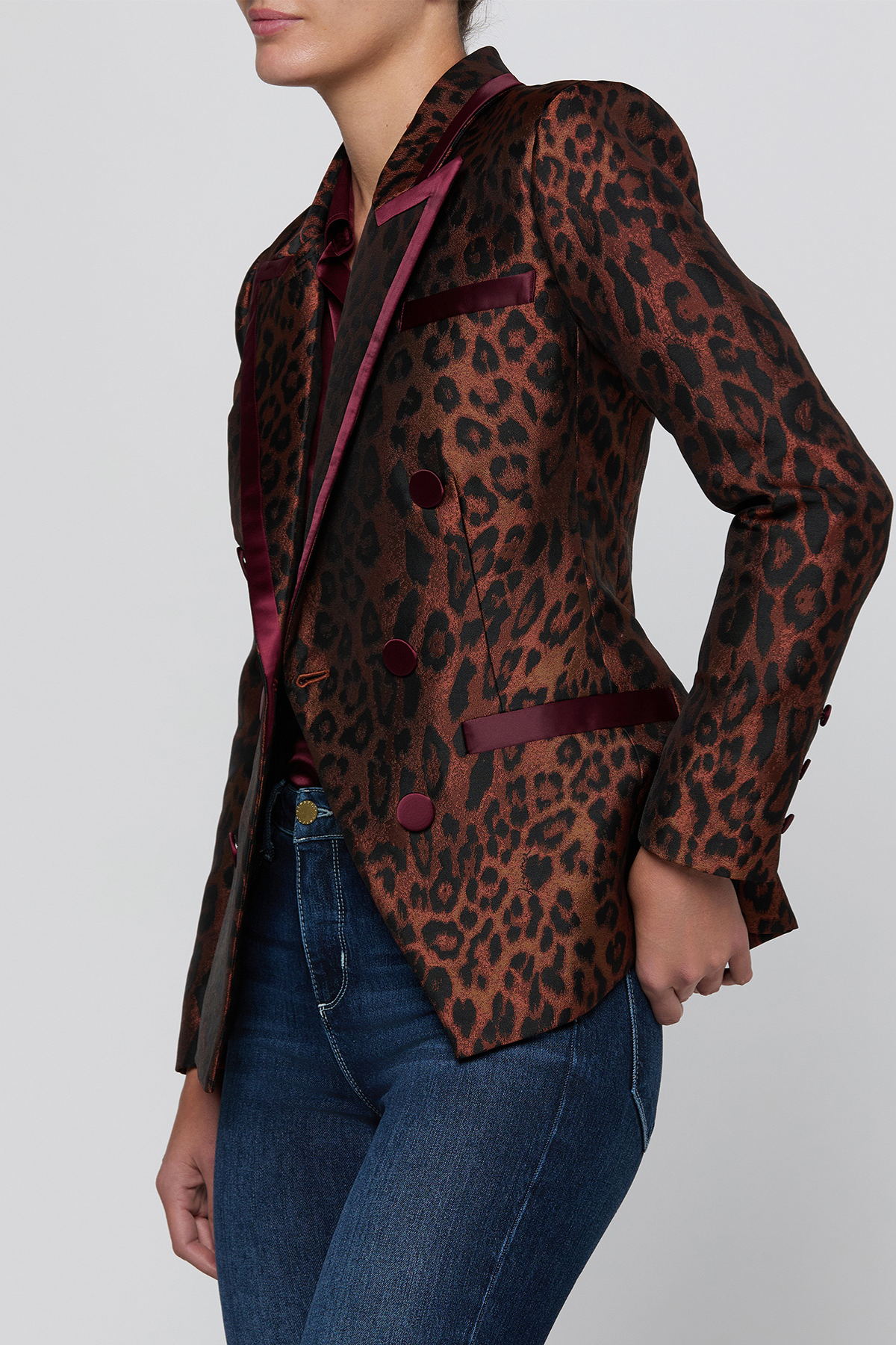 A woman wearing a fitted leopard-print blazer with dark burgundy satin lapels and buttons, paired with blue jeans, photographed against a light gray studio background.
