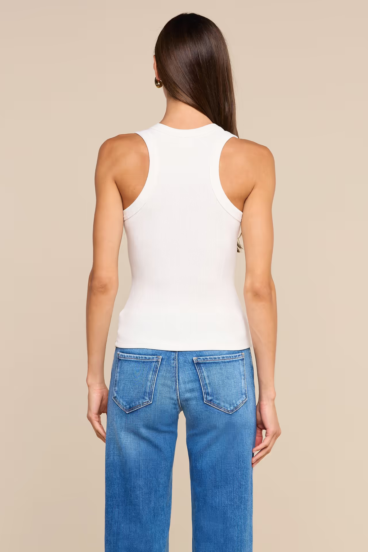A woman shown from the back wearing a fitted white ribbed tank top and blue jeans against a plain beige studio background.