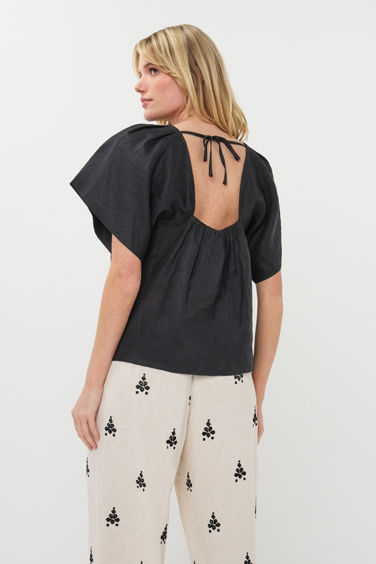 Woman wearing a black top with open back and white pants with black patterns on a white background